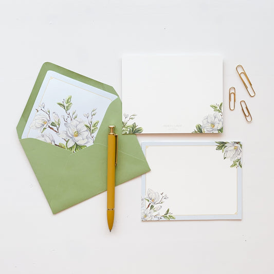 White stationery card with pale blue pin stripe border featuring white magnolia watercolor artwork. Light green envelope lined with matching magnolia watercolor artwork.