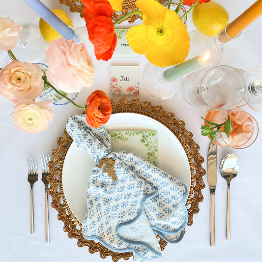 Styled table top with silver flatware, rattan charger, white ceramic plate, acrylic menu, white and blue bastik patterned cloth napkins tied with laser cut wooden charm in the shape of a maidenhair fern. Lit colorful candles, bright poppies and ranunculus and acrylic place cards.